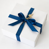 The Sun and Sand Gift Box is a white box tied with a wide, dark blue satin ribbon and bow, adorned with a small dried flower on top, set against a plain white background.