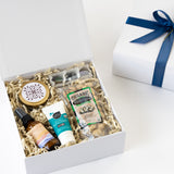 The Sun and Sand Gift Box features a blue ribbon and includes a round travel candle tin, spray bottle, mineral sport sunscreen, organic pistachios, and chocolates, all nestled in crinkle paper.