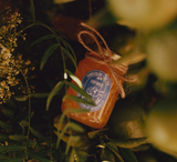 A glass jar of Sunrise Pixie Tangerine Marmalade, adorned with a blue label and twine bow, rests among lush green leaves and foliage in gentle, natural light.