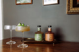 Two crystal coupe glasses, one garnished with cucumber slices, sit beside two bottles of Tandem Smoky & Oaky Non-Alcoholic Cocktail on a brass tray atop a wooden table—ready to serve against a gray wall with framed artwork.