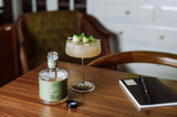 A glass of Tilden Non-Alcoholic Cocktail - Lacewing with cucumber ribbons sits on a wooden table beside an open Tilden bottle and a closed notebook with a pen, with a chair and drawers visible in the background.