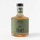 A clear glass bottle labeled Tilden Non-Alcoholic Cocktail - Lacewing contains a golden, herbaceous, ready-to-serve drink. Its green label highlights it as a fresh, 0% alcohol social beverage with a black cap.