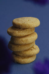 A stack of six golden Verdon Lavender Shortbread Cookies, made in California, is neatly arranged on a blue surface, with blurred lavender floral shapes in the foreground.