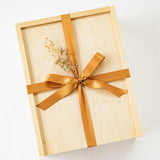 The À Votre Santé Gift Box is a light wooden box tied with a wide golden satin ribbon and bow, accented by a small sprig of dried flowers, resting on a white background—perfect for virtual event gifting.