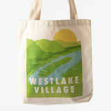 The Westlake Village Canvas Tote is a sustainable bag made from recycled cotton, featuring thick handles and a graphic of green hills, a river, and a yellow sun above bold white “WESTLAKE VILLAGE” text.