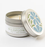 A small silver tin of Wildflower Travel Candle with hand-poured soy wax, labeled Santa Barbara Company. The lid, partially off to reveal the candle, displays a blue flower and green leaves illustration.