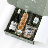 The Wine & Dine Gourmet Gift Box includes Sauvignon Blanc, dried orange slices, organic pistachios, seasoned almonds, olives, and chocolates—arranged on green shredded paper in a green box.