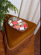 A clear tray of scarlet red Matchbox Merry Christmas boxes on a triangular fringed stool.