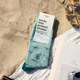 Socks that Protect Whales in Conscious Step packaging on a beach towel in the sand beside an open book