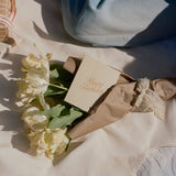 A bouquet of white tulips in brown paper with a lace ribbon and an embossed Happy Birthday Note Card tucked inside.