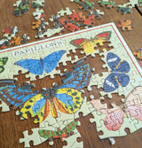 A partially completed butterfly-themed jigsaw puzzle sits on a wooden table, with scattered, unplaced pieces around the edges. The puzzle features colorful butterflies and the word PAPILLONS at the top.