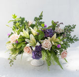 Soft and dreamy floral compote with blush roses, cream lilies, purple hydrangea, purple filler and mixed greenery in a white pedestal bowl.