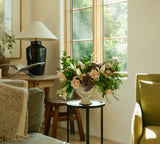 A Centerpiece Floral Arrangement brightens a small table by a sunny window in a cozy, softly lit living room.