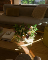 A Centerpiece Floral Arrangement sits on a coffee table in a sunlit living room near a sofa and books.