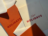 Two Happy Holidays Note Cards, one orange and one cream, each embossed with the words Happy HOLIDAYS in red.