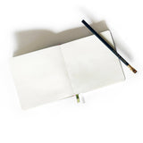 A Square Linen Blank Notebook lies open with a black pencil across its pages on a white background.