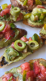 Toasted bread topped with green olives, diced tomatoes, and small-batch Zucchini Pesto on a white surface.