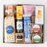 A curated gift box filled with assorted snacks and treats, including a large bag of Hatch's popcorn, a jar of artisanal caramel, Salami, cookies, dried apricots, and other packaged foods, arranged in a light green box on a white background.