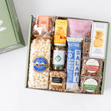 A curated gourmet gift box filled with assorted snacks and treats, including popcorn, cookies, crackers, nuts, and packaged food items, displayed in a light green box on a white background.