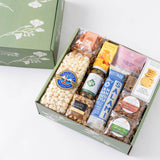 A gourmet gift hamper box filled with assorted snacks and treats, shown open with its matching green floral lid beside it on a white background. 