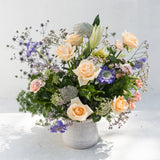 An elegant floral arrangement featuring peach roses, purple stock, baby's breath, and green foliage in a textured gray ceramic pot, set against a clean white background.
