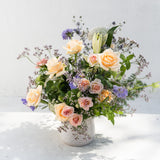 A vibrant flower bouquet in a textured white ceramic pot, featuring peach and cream roses, purple flowers, and green foliage, set against a clean white background.