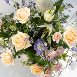 A top-down photo of a floral arrangement featuring pale peach and cream roses, a cream tulip, purple anemone flowers, and green foliage on a light gray background.