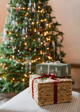 Two wrapped holiday gifts, one in a woven basket with a red ribbon and the other in a green box, sit in front of a decorated Christmas tree with lights and tinsel.