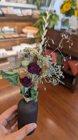 A hand holds the California Dried Floral Arrangement with organic blooms and purple roses in a cozy, wood-floored shop.