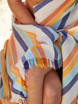 A close-up of a striped beach towel or wrap with fringed edges, draped over a person's lap. The fabric features diagonal stripes in white, navy blue, orange, and light blue.
