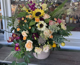 The Regal: A seasonal flower arrangement with sunflowers, roses, and greenery in a stone vase on wood.