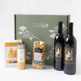 A green Santa Barbara Company mailer gift box with a poppy design, beside a box of Rustic Bakery cheese coins, a tin of caramelized pear tea, a bag of vanilla salted caramel corn, and two bottles of Theopolis Vineyards red wine.