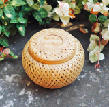 The Petite Woven Basket, handmade with a lid, sits on stone beside leafy plants.