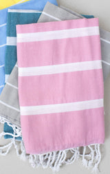 A stack of folded Baby Hammam Towels in pastel pink, blue, teal, yellow, and gray, each featuring white stripes and tasseled fringes at the ends.