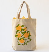 The California Oranges Organic Cotton Tote is a beige canvas bag with handles, decorated with California oranges, green leaves, white blossoms, and “california” in script at the bottom. Yellow paper peeks from the top.