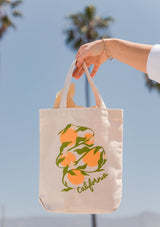 A hand holds the California Oranges Organic Cotton Tote, featuring an illustration of oranges, green leaves, white flowers, and "California" text. Palm trees and a clear blue sky create a vibrant made-in-USA backdrop.