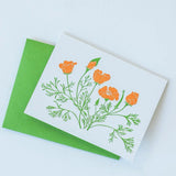 The California Poppy Letterpress Greeting Card features an illustration of orange poppies and green stems on a white card atop a bright green envelope, capturing the vibrant spirit of Santa Barbara.