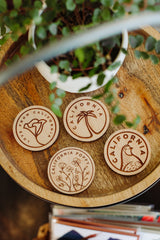 The California Quail Wood Magnet, crafted from sustainably harvested wood and featuring minimalist illustrations, is displayed on a tray next to a potted plant.