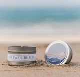 A tin candle labeled Miramar Beach sits on sandy beach, its lid beside it showing waves and a sunset; the blurred ocean and sky in the background evoke the relaxing vibe of the California Scene Gift Box.