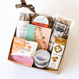 The California Scene Gift Box is filled with assorted snacks and treats like wild berry mix, sea salt caramels, dried figs, a seed & nut bar, organic pistachios, a scented candle, a cinnamon treat, and a colorful California-themed card.