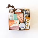 The California Scene Gift Box features organic pistachios, wild berry mix, a macadamia seed & nut bar, caramel popcorn, honey cinnamon almonds, chocolate, a small tin, and a California map coaster—all tied with a brown ribbon.