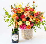 Champagne + Flowers: Sustainably grown orange, yellow, and pink orchids in a white textured vase are paired with a bottle of French Champagne, beautifully displayed on a white surface.