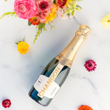 A bottle of Chandon Brut Sparkling Wine - 375ml rests on white marble, surrounded by colorful flowers and a bouquet of pink, yellow, and orange blooms, creating a vibrant scene that showcases this California sparkling wine.