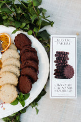 A plate of buttery Chocolate Cacao Nib Shortbread Cookies, made with organic wheat flour, sits beside the box, garnished with mint leaves and a dried orange slice.