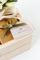 A Chocolate Gift Box with a green ribbon and "Jessica Foster Confections" label is placed in a wooden crate, featuring handcrafted chocolate truffles and white flowers partially visible at the top.