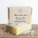 Two rectangular bars of Dead Sea Minerals Organic Soap rest on a wooden surface with coarse sea salt scattered around them. The label highlights organic, palm-oil free ingredients—ideal for sensitive skin.