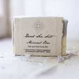 A rectangular handmade soap bar labeled Dead Sea Minerals Organic Soap sits on a light surface with scattered salt crystals. The label highlights that it’s palm-oil free, organic, vegan, and ideal for sensitive skin with no chemical preservatives.