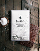 A Dick Taylor Fleur de Sel Chocolate bar with an illustrated sailboat wrapper sits on a rustic wooden surface next to an unwrapped bar and a small bowl of Guatemalan sea salt.