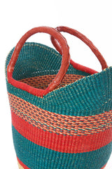 The Fair Trade Woven Basket is crafted from elephant grass with red, orange, and teal stripes and two brown leather-wrapped handles. This Ghanaian shopper is pictured against a white background.