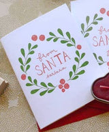 The From Santa (Barbara) Christmas Note Cards feature a green and red wreath with berries and "from SANTA Barbara" in the center, displayed beside a red candle and envelope on a patterned surface.